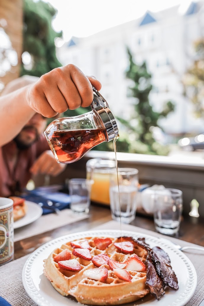 Drizzling syrup onto a belgian waffle with strawberries and bacon